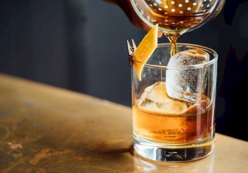 A glass of whiskey on the rocks being poured, with an orange twist garnish resting on the rim, on a wooden bar surface.