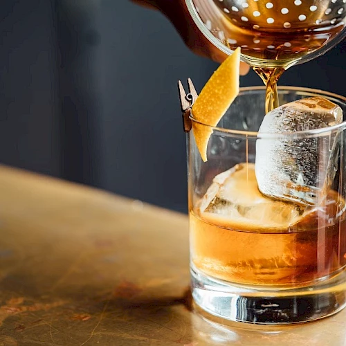 A glass of whiskey on the rocks being poured, with an orange twist garnish resting on the rim, on a wooden bar surface.