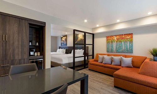 A modern, open-plan living space with a glass partition between living and bedroom, orange sectional sofa, a dark dining table, and warm artwork on the wall.