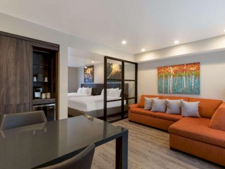 A modern studio with a separate sleeping area, featuring a glass partition, dark wood cabinetry, a black dining table, and an orange sectional sofa with gray cushions.