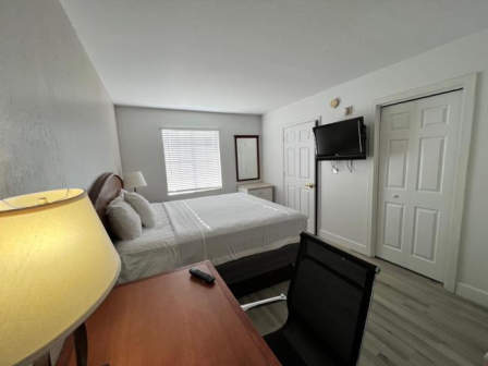 A small hotel-style bedroom with a bed, desk, chair, wall-mounted TV, and a window letting in light, plus a dresser and mirrored door closet.