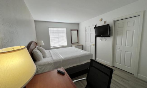 A small hotel-style bedroom with a bed, desk and chair, wall-mounted TV, dresser, and a window letting in soft light, door and closet doors nearby.