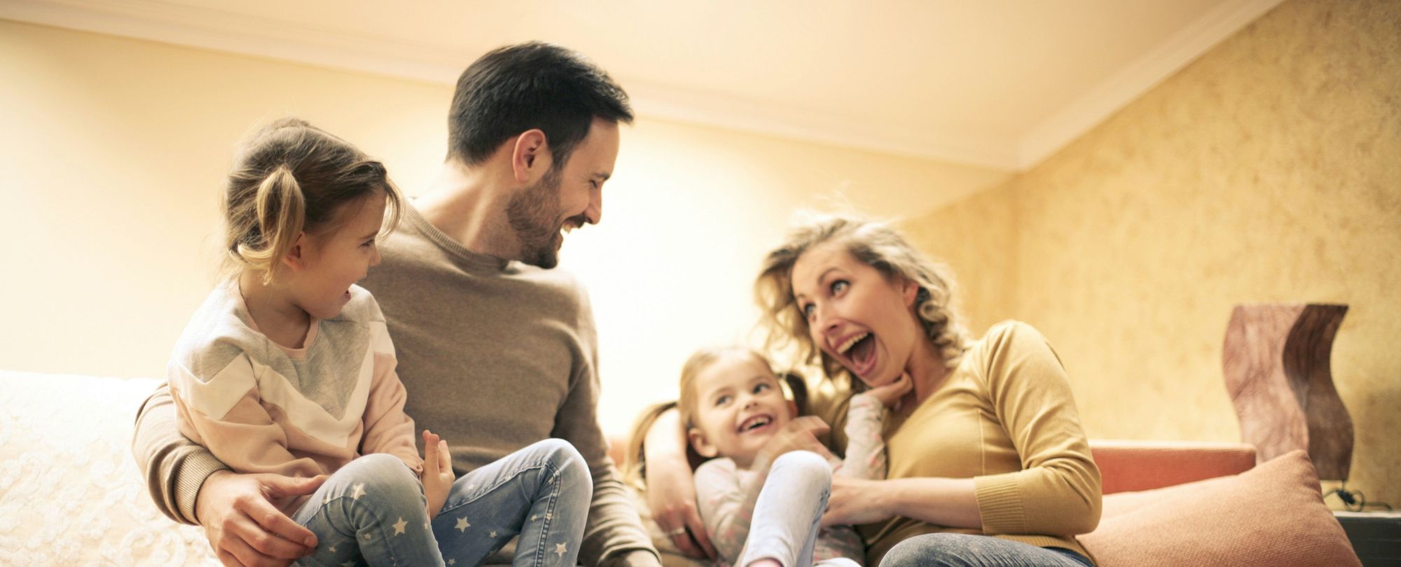 A happy family sits on a sofa, kids giggling with parents, cozy living room, mugs on the table, warm lighting.