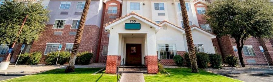 A residential apartment building with a light-colored facade, two tall palm trees, a well-kept lawn, and a bright blue sky.