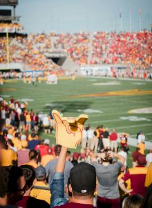Cheering fans fill a football stadium, a hand sign points to the field as the game unfolds beneath a bright, crowded stands.