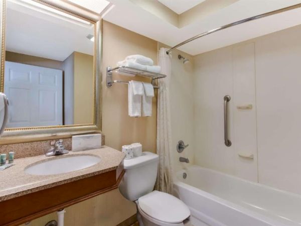 A compact hotel bathroom with a beige theme: sink with marble counter, mirrored frame, toilet, and a white bathtub/shower combo with grab bar; towel rack above the toilet.