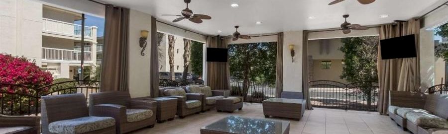 A spacious, outdoor lounge area with wicker sectional seating, a central coffee table, ceiling fans, and tiled flooring, perfect for relaxing.