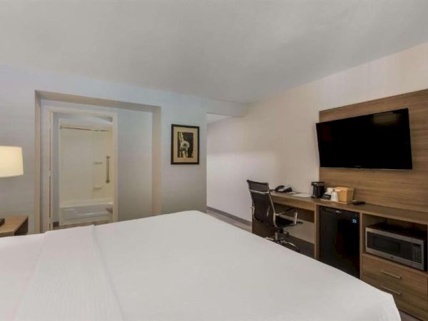 A modern hotel room with a bed in foreground, desk and wall-mounted TV, mini-fridge, and a doorway leading to a small bath area.