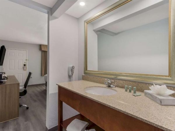A hotel bathroom with a large framed mirror and a granite counter, plus a small workspace with a chair and desk in the next room, neat and modern.