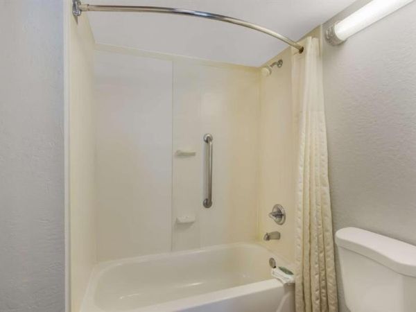 A small bathroom with a white bathtub and shower, curved shower curtain rod, beige curtain, and a white toilet visible; plain light walls.