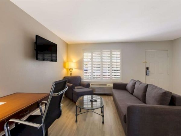A compact living room with a gray sectional, glass coffee table, wall-mounted TV, desk and chair, and a lit lamp by a window with blinds, ending here.