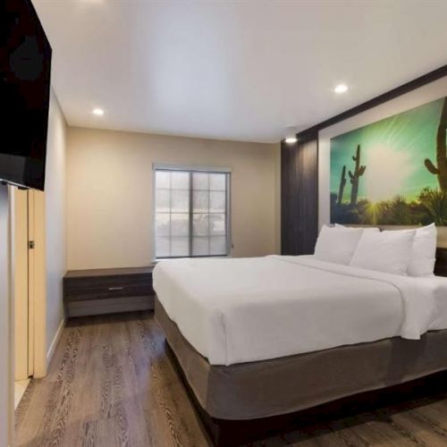 A modern hotel room with two queen beds, dark wood accents, a large cactus landscape artwork, a wall-mounted TV, and a small desk area, bright and clean.