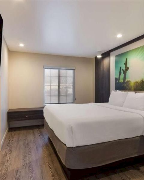 A modern hotel room with two queen beds, dark wood accents, a large cactus landscape artwork, a wall-mounted TV, and a small desk area, bright and clean.
