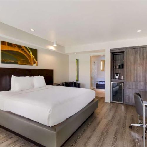 A modern hotel room with a large bed, wooden headboard, neutral decor, a desk with chair, a wall-mounted TV, wardrobe, mini-fridge, and a doorway to a bathroom.