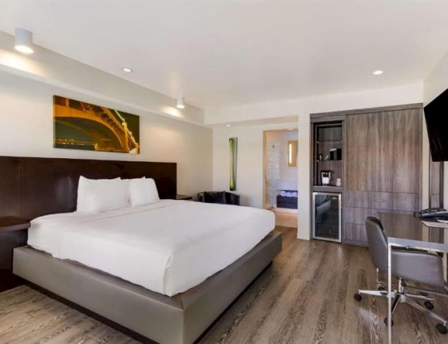 A modern hotel room with a large bed, wooden headboard, neutral decor, a desk with chair, a wall-mounted TV, wardrobe, mini-fridge, and a doorway to a bathroom.