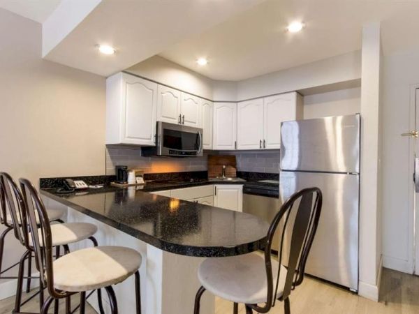 A modern kitchen with white cabinets, black countertops, stainless steel fridge and appliances, an island with bar stools, and recessed lighting.