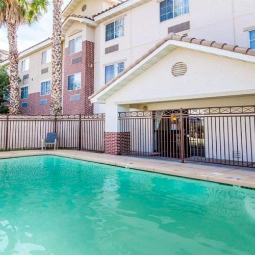 A apartment complex with a fenced outdoor swimming pool and a covered patio area, light-colored buildings, and palm trees.