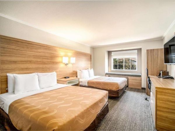 A hotel room with two double beds, wooden headboards, lamps, a window, desk, mini-fridge, and TV, tidy and warmly lit.