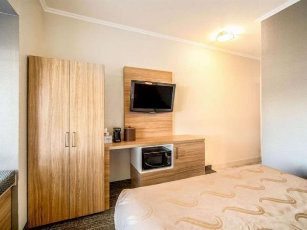 A tidy hotel room with a wooden wardrobe, a wall-mounted flat-screen TV, a small desk with a microwave below, and a neatly made bed featuring a patterned beige bedspread.