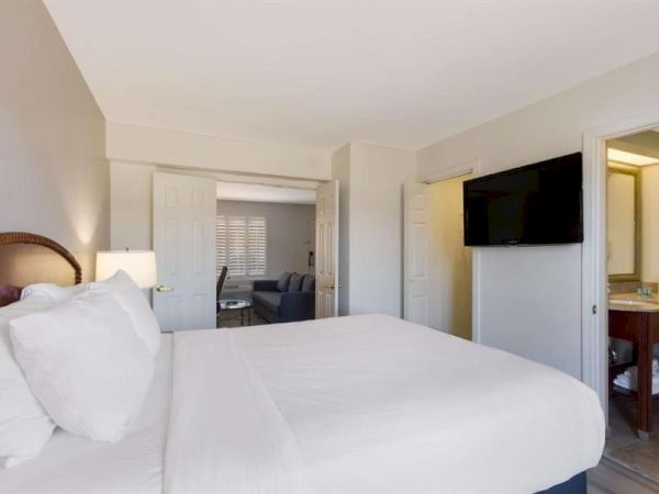 A clean hotel room with a large bed, wall-mounted TV, open bathroom area, and a seating nook visible in the doorway, modern and bright.