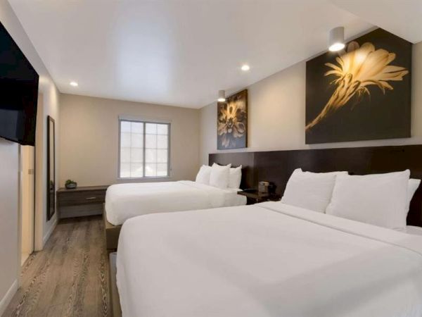 A modern hotel room with two queen beds, white linens, a dark wood headboard, wall art, a window, and a small desk area on the far wall.