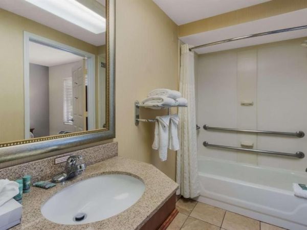 A hotel bathroom with a single sink, granite countertop, large mirror, and a bathtub/shower combo with grab bars and towels, plus a beige-tiled floor.