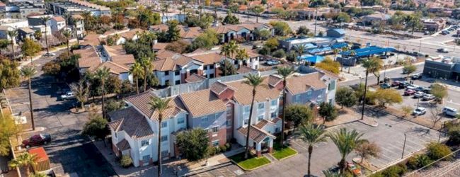 An aerial view of a suburban neighborhood with streets, houses, palm trees, parked cars, and a few apartment buildings under clear skies.