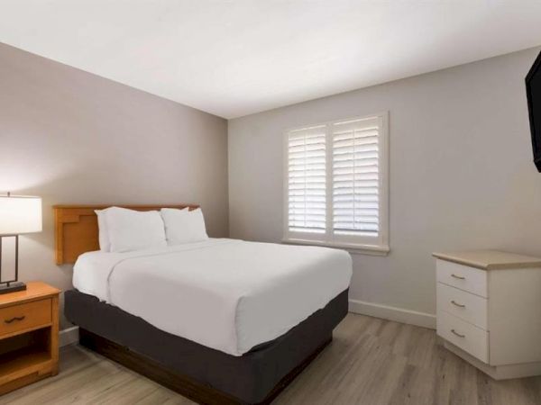 A simple hotel-style bedroom with a queen bed, nightstand lamp, white bedding, a dresser, and a window with shutters, neutral tones throughout.