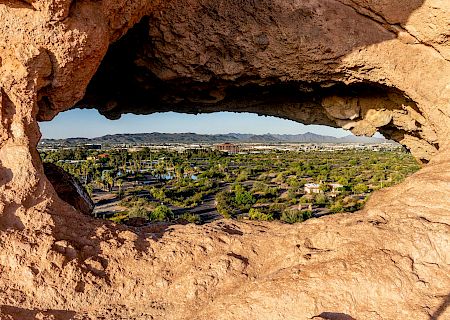 A narrow natural rock arch with a small opening showing a blue sky and distant landscape, formed by eroded sandstone sandstone framing a tiny view.