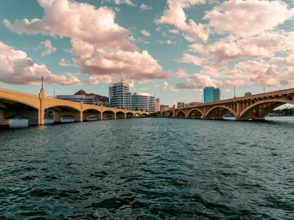 A city river scene with a long bridge across calm water, modern buildings in the background and a partly cloudy sky overhead, sunny and vibrant.