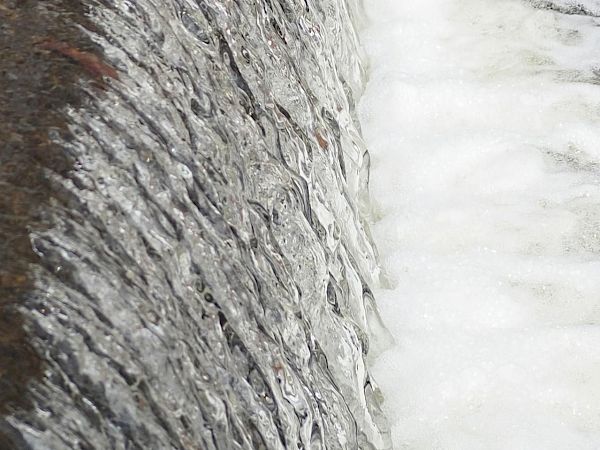 A rough stone or concrete wall next to fast-flowing white water, like a dam or spillway edge.