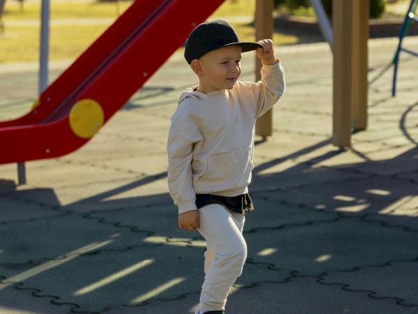 A young boy on a skateboard at a playground, wearing a cap and white outfit, standing near a red slide with a sunny backdrop.